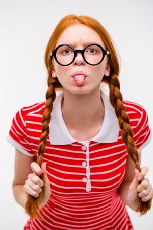 Redhead funny amusing girl with two braids in red striped t-shirt and round glasses showing tongue posing on white backgroundの写真素材