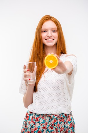 Cheerful candid happy pretty redhead girl in white top and floral skirt holding empty glass and offering ripe orangeの写真素材