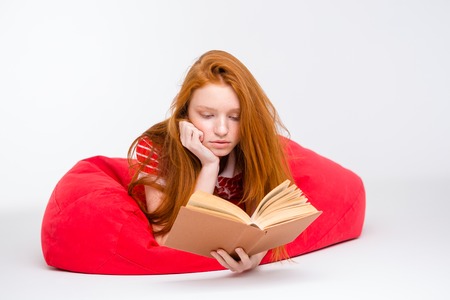 Attractive redhead concentrated girl lying and relaxing in red bean bag and reading a book isolated on white bakgroundの写真素材
