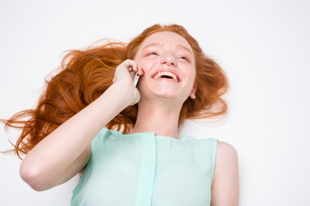 Cheerful happy girl with beautiful long hair lying and talking on mobile phone over white backgroundの写真素材