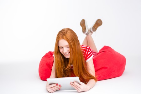 Sensual cute smiling young lady with beautiful long red hair lying on red bean bag using tablet on white backgroundの写真素材