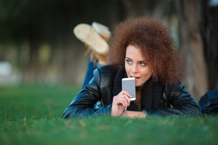 Portrait of a thoughtful woman with curly hair lying on green grass with smartphone outdoorsの写真素材