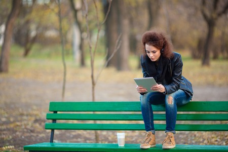 Portrait of a casual woman sitting on the bench and using tablet computer in autumn parkの写真素材