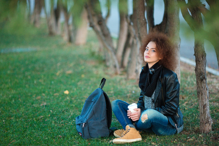 Portrait of a young female student sitting on green grass with cup of coffee and backpack outdoorsの写真素材
