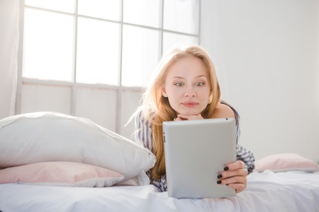 Surprised wondered blonde young woman lying in bed and using tablet PCの写真素材