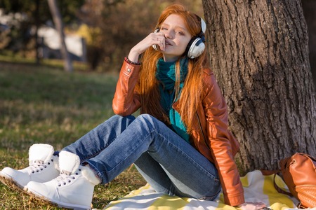 Pretty pensive young woman in leather jacket, white boots and jeans listening to music under the treeの写真素材