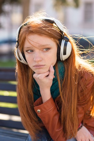 Portrait of beautiful sad pensive young woman with long red hair in headphones sitting in the parkの写真素材
