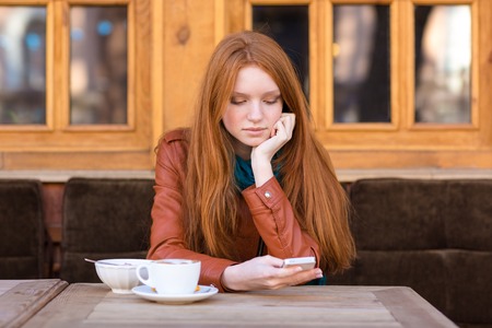 Pretty thoughtful girl with beautiful long red hair in leather jacket using smartphone and drinking coffee in outdoor cafeの写真素材