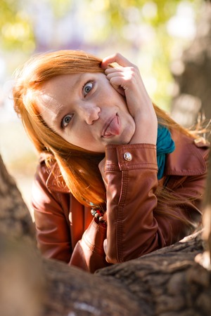 Pretty amusing redhead girl making funny face and showing tongue posing in park near treeの写真素材