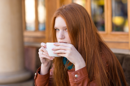 Attractive thoughtful cute girl with long beautiful red hair drinking coffee in outdoor cafeの写真素材
