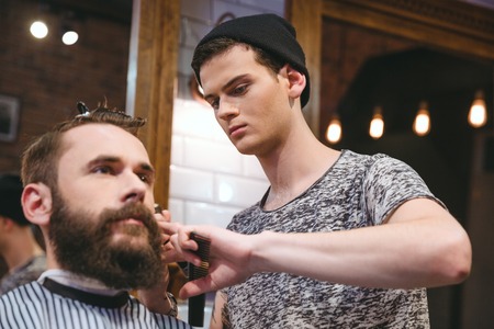 Young skillful barber making haircut to handsome bearded man in barbershopの写真素材