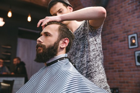 Young handsome barber cutting hair of young attractive bearded hipster in barbershopの写真素材