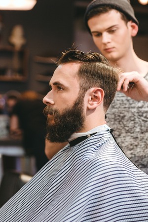 Professional hairdresser cutting young bearded man's hair in hairdressing salonの写真素材