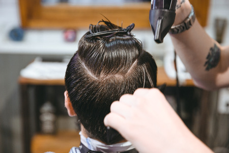 Closeup of hands of hairstylist drying hair of male client using hairdryer and comb in barbershopの写真素材