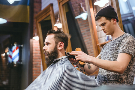 Barber making haircut to handsome attractive man with beard and working with electric razorの写真素材