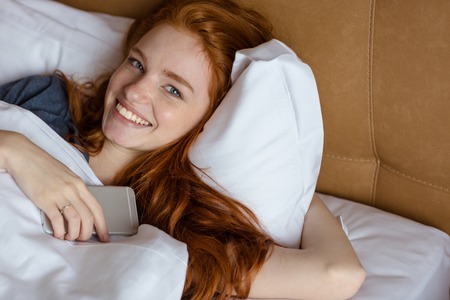 Portrait of a smiling redhead woman lying in the bed with smartphone and looking at cameraの写真素材