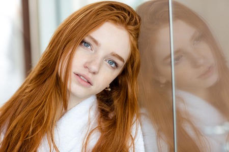 Portrait of a beautiful redhead woman in bathrobe leaning on wall and looking at  cameraの写真素材