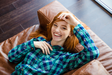 Beautiful joyful natural young woman in checkered shirt laying on brown leather sofa and laughingの写真素材