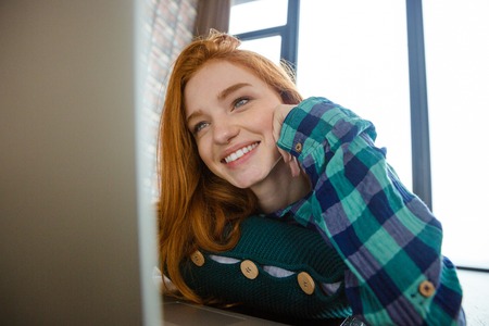 Joyful lovely cute young woman with red hair in checkered shirt watching on monitor of laptop and...