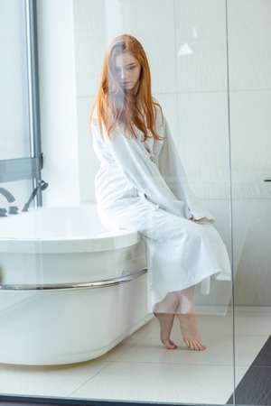Portrait of a young redhead woman in bathrobe sitting in bathroomの写真素材