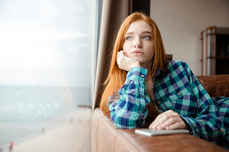 Dreaming thoughtful lovely cute girl with long red hair in checkered shirt looking out of the window and listening to musicの写真素材