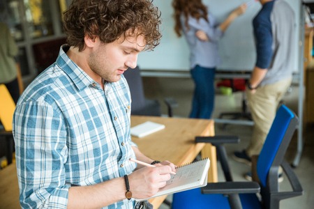 Thoughtful confident young curly man in blue plaid shirt making notes in notebook and creating project with colleaguesの写真素材