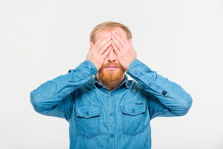 Portrait of young handsome blond bearded male in jeans shirt with eyes covered by hands over white backgroundの写真素材