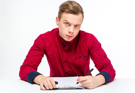 Suspicious curious attractive young male in red shirt with raised eyebrow writing on clipboard over white backgroundの写真素材