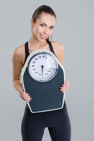Portrait of happy lovely cute fitness girl in sportwear with weigh scale over grey backgroundの写真素材