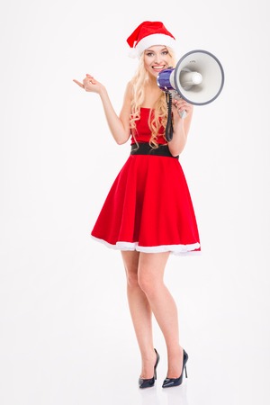 Full length of joyful attractive blonde woman in santa claus dress and hat with megaphone pointing away over white backgroundの写真素材