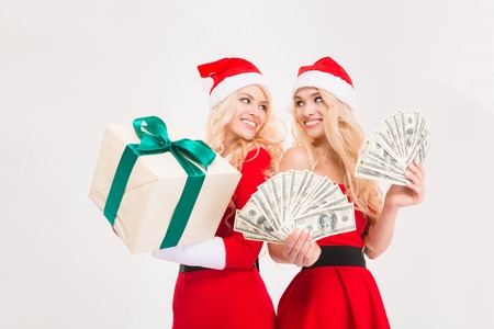 Beautiful happy sisters twins in red santa claus clothes and hats  with money and gift isolated over white backgroundの写真素材