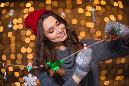 Pretty cute smiling girl in grey sweater and mittens making handmade christmas decoration over holidays lights backgroundの写真素材