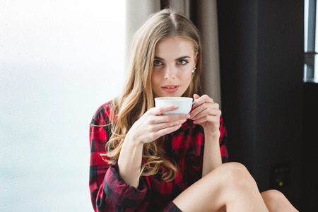 Beautiful thoughtful young woman in plaid shirt drinking coffee in the morningの写真素材