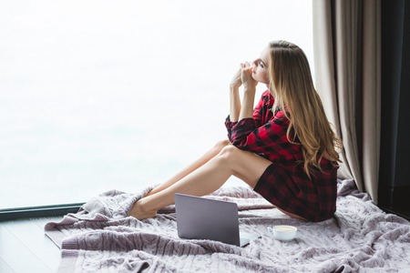 Attractive thoughtful young female in checkered shirt sitting on the floor and looking through the windowの写真素材