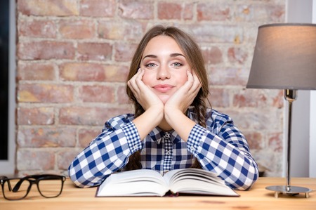 Portrait of a cute female student sitting at the table with book and looking at camera