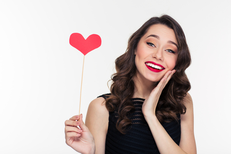 Happy pretty shy young female with retro hairstyle holding heart props isolated over white backgroundの写真素材