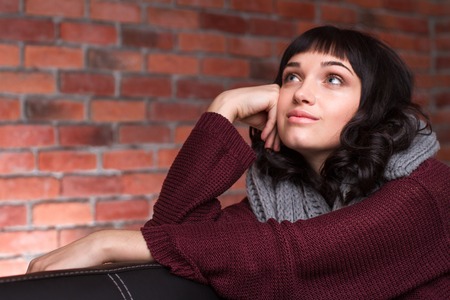 Lovely cute young woman in knitted clothes sitting and dreaming over brick wall backgroundの写真素材