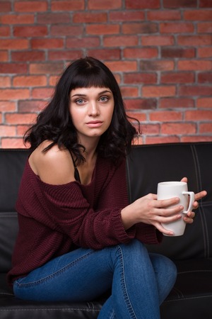 Charming thoughtful young woman in sweater and jeans sitting on couch and drinking tea over brick wall backgroundの写真素材