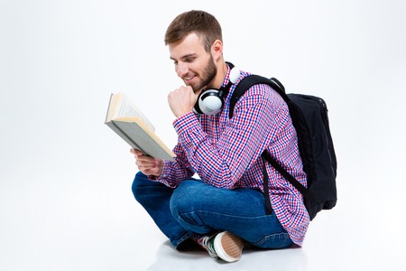 Cheerful handsome bearded young man in checkered shirt with backpack and headphones reading book and...