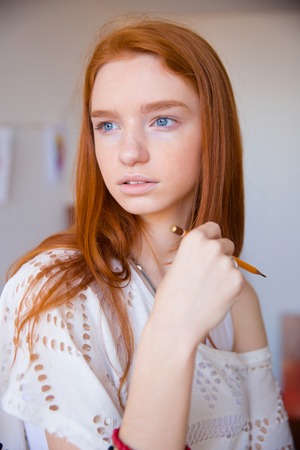 Portrait of beautiful thoughtful young woman with long red hair holding pencil and thinkingの写真素材