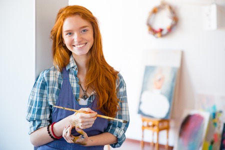 Cheerful beautiful young woman painter with long red hair holding brush and standing in art studioの写真素材