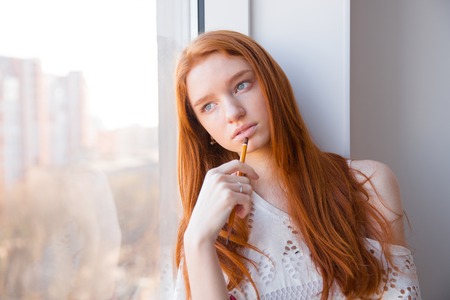 Beautiful thoughtful young redhead woman with pencil dreaming near windowの写真素材