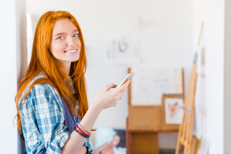 Charming happy young woman with long red hair using smartphone in art workshopの写真素材