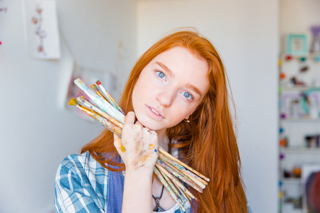 Beautiful pensive young woman painter with long red hair holding paintbrushes in art studioの写真素材