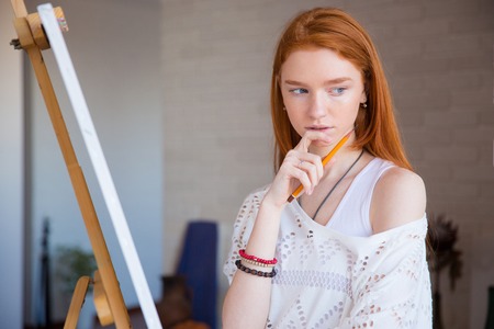 Pensive attractive young female artist with red hair standing and thinking near easel in drawing classの写真素材