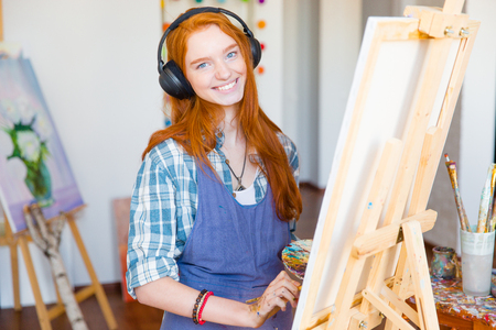 Charming smiling young woman artist in headphones and apron painting on canvas and listening to music in art studioの写真素材