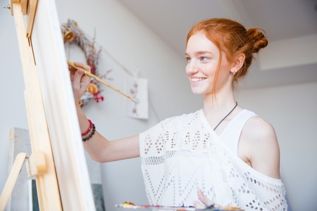 Smiling attractive young woman painter with red hair painting on canvas in artist workshopの写真素材