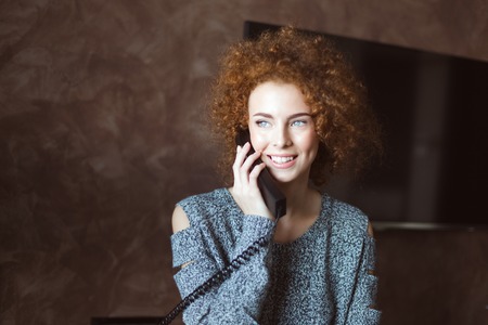 Beautiful happy curly woman in grey knitted sweater talking on landline telephone in the roomの写真素材