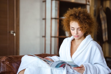 Cute lovely serene redhead young female sitting at home and reading magazineの写真素材