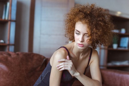Alluring sensual young curly redhead woman sitting on sofa and thinkingの写真素材